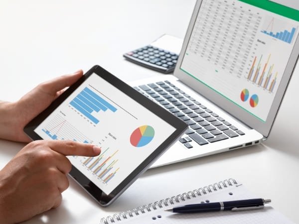 Une personne consulte des graphiques et des tableaux de données sur une tablette et un ordinateur portable posés sur un bureau.